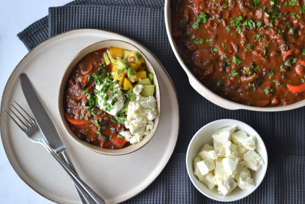 A bowl of black bean chilli with feta and mango salsa, sitting on a plate with cutlery and a black napkin. There is a pot of chilli and feta to the right of the image.